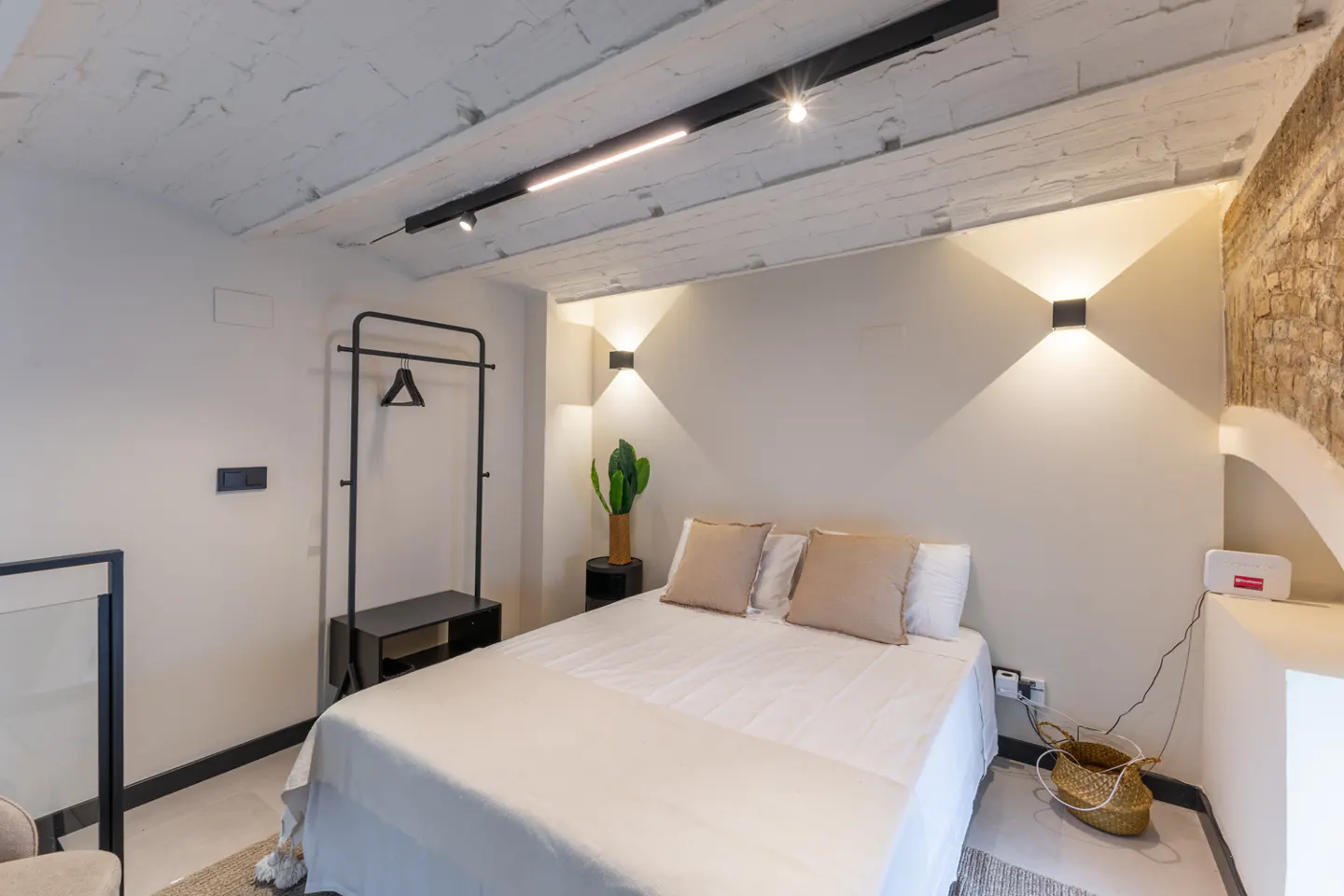Bedroom with white walls, a bed with beige pillows, and a black clothing rack. The ceiling has exposed beams and modern lighting.