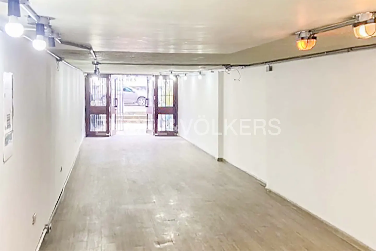 Empty commercial space with white walls, wood floors, and open double doors to the street. Track lighting illuminates the room.