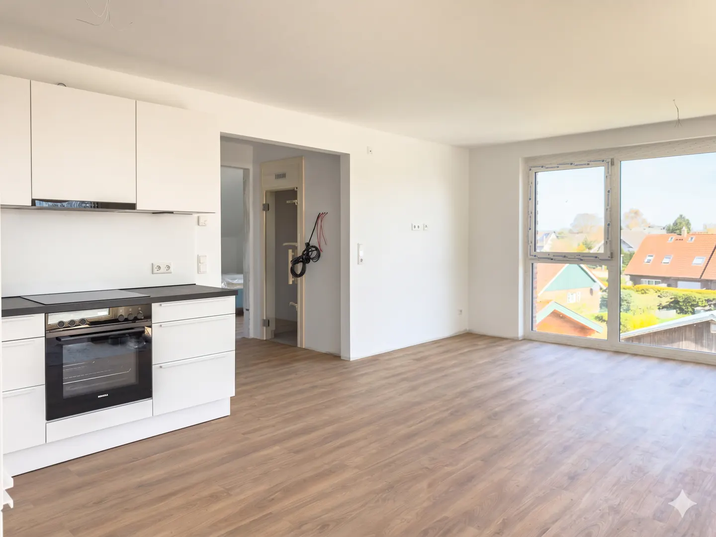Bright, empty apartment with wood floors, white walls, and a modern white kitchen. A large window overlooks houses with red roofs.