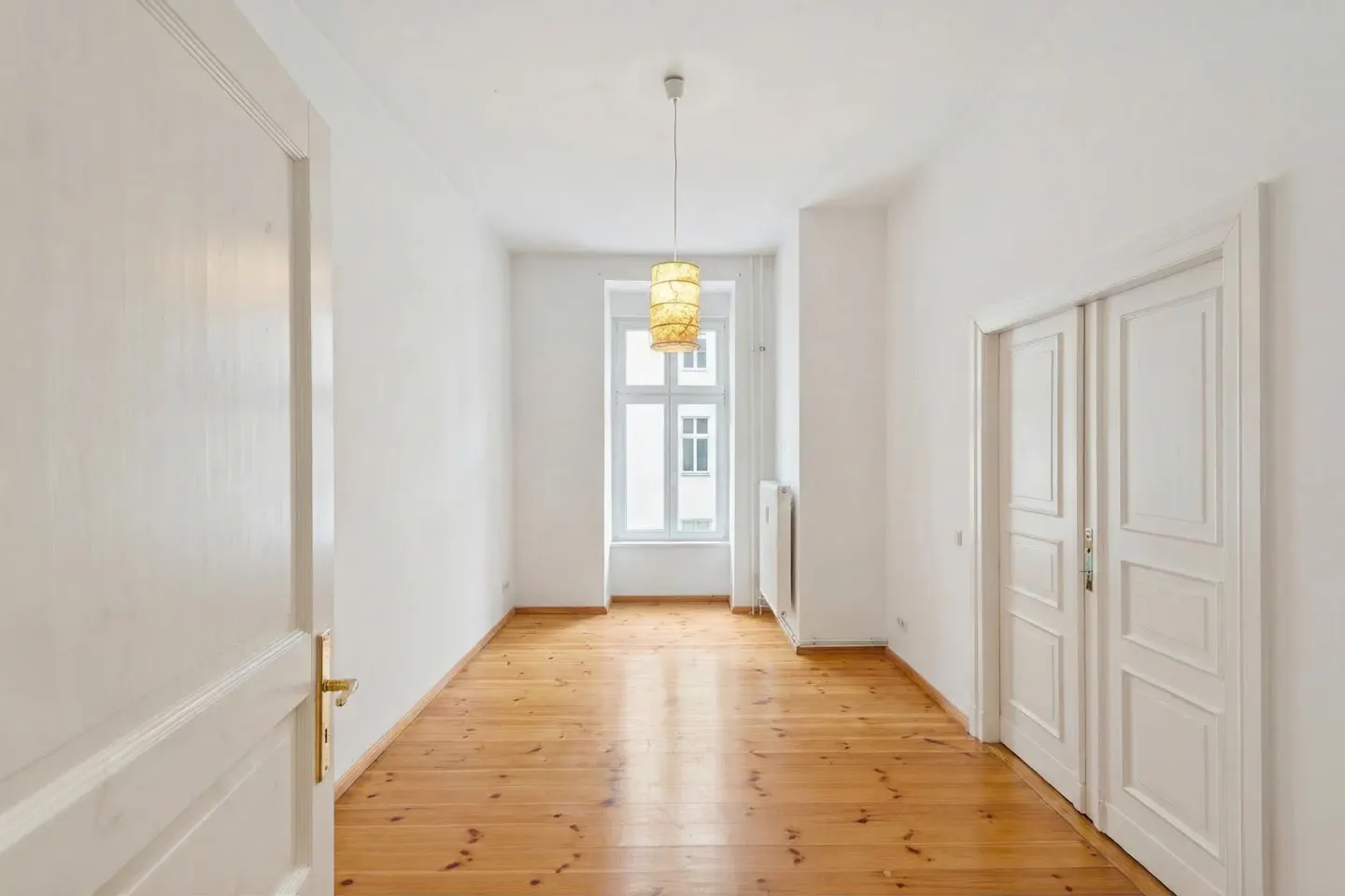 Bright, empty room with wood floors, white walls, and a window. A pendant light hangs from the ceiling. A white door is ajar on the left.