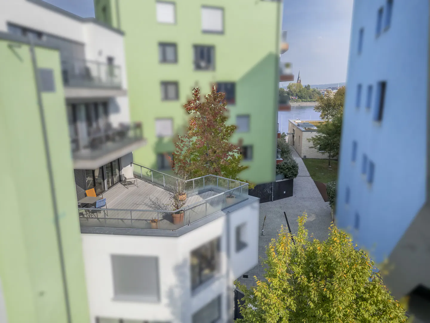 View of a modern apartment complex with green and blue buildings, a balcony with furniture, and a river in the background.