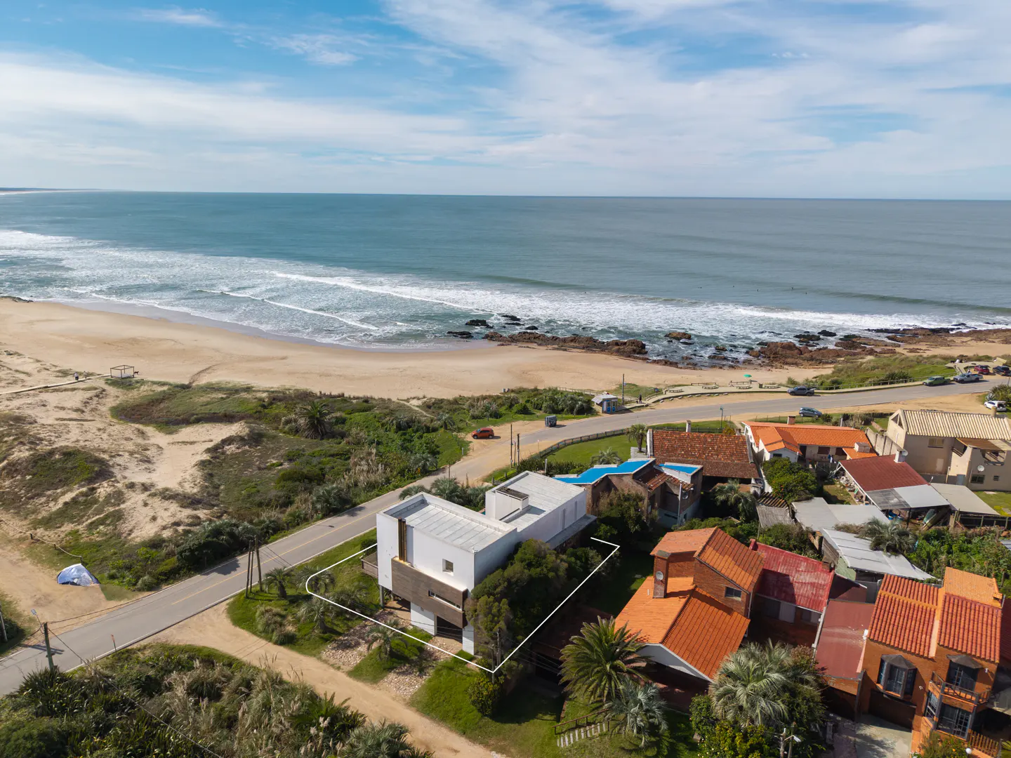 Aerial view of a modern white house with a brown accent wall, near a sandy beach and blue ocean under a cloudy sky.