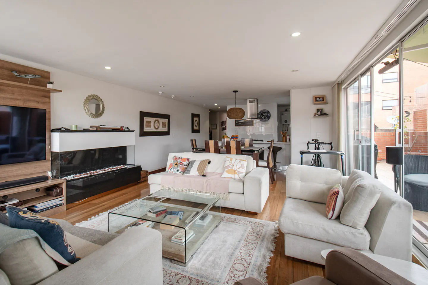 Bright, open-concept living room with white sofas, glass coffee table, and a modern fireplace. Dining area and kitchen visible in the background.
