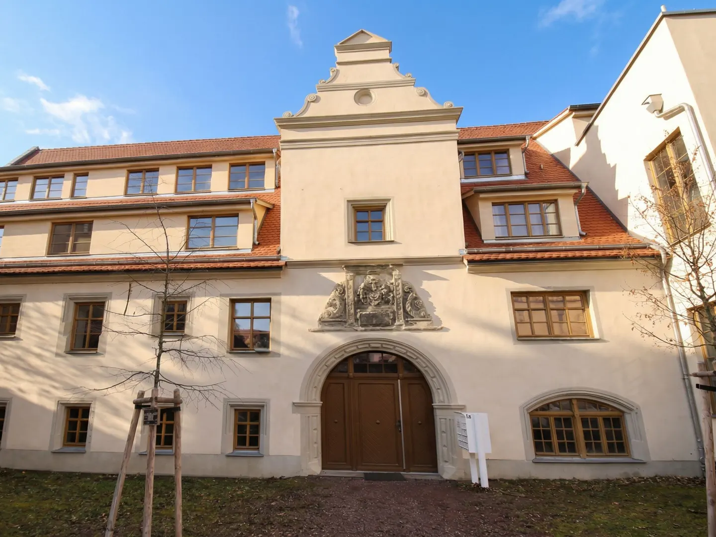 Exterior view of a three-story building with a red tile roof and light yellow walls, featuring an arched entrance with brown doors.
