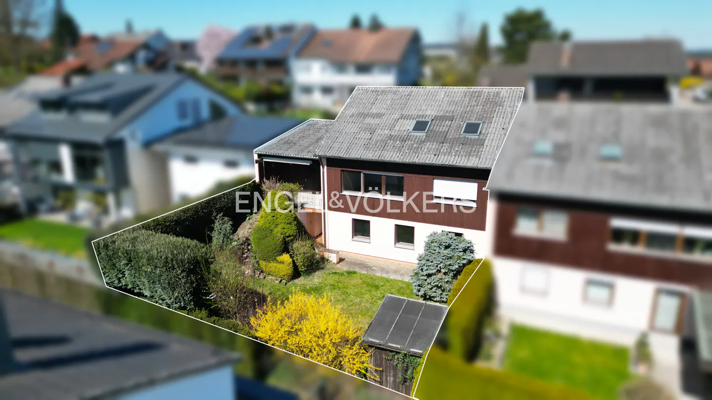 Two-story house with a gray roof, brown and white siding, and a green lawn. Bushes and yellow flowers are in the yard. Engel & Volkers logo.