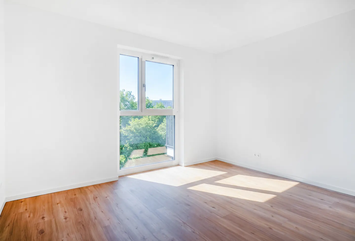 Bright, empty room with white walls and wood floor. Sunlight streams through a window with a view of green trees.