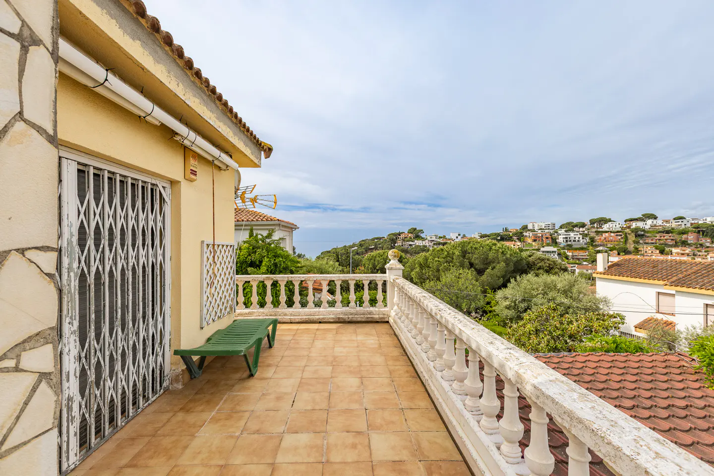 Balcony view with tan tile floor, white balustrade, and green bench. Rolling hills and houses in the background.