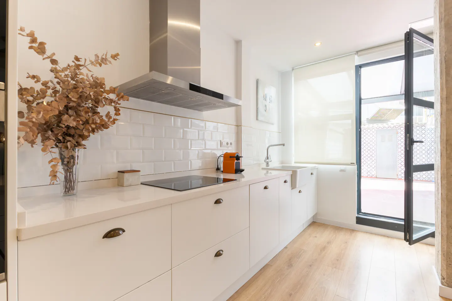 Bright kitchen with white cabinets, subway tile, and wood floors. Stainless steel range hood and open door to the outside.