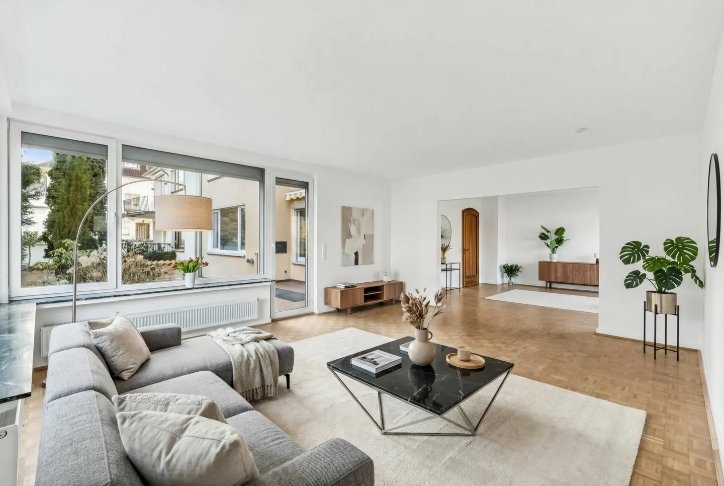 Bright living room with a gray sectional sofa, black marble coffee table, and large windows overlooking a garden.