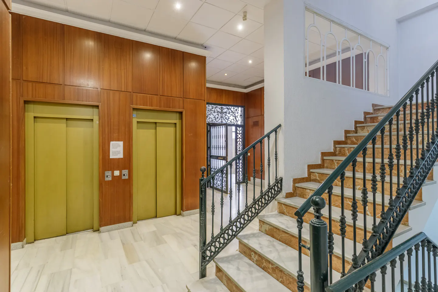 Interior view of a building lobby with two olive-green elevators, marble stairs with black railings, and a decorative metal gate.