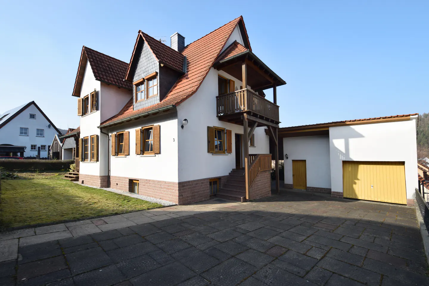 Two-story white house with brown shutters, a red-tiled roof, and a wooden balcony, next to a white garage with a yellow door.