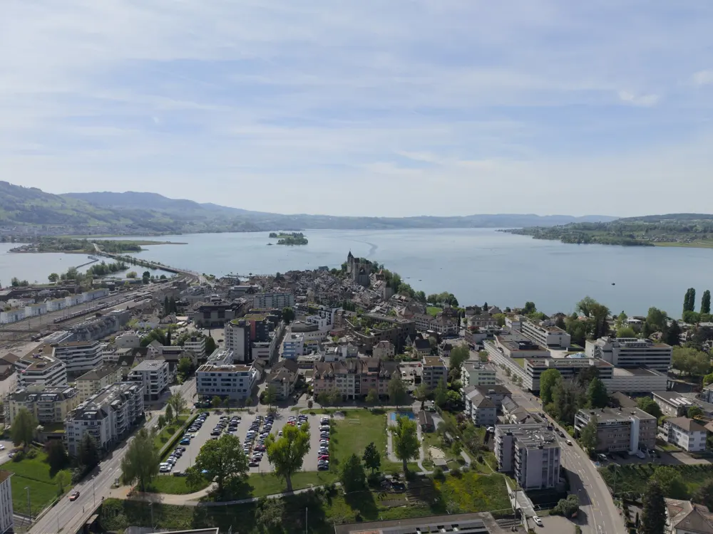 Aerial view of a city with buildings, a parking lot, and a lake with mountains in the background under a blue sky.