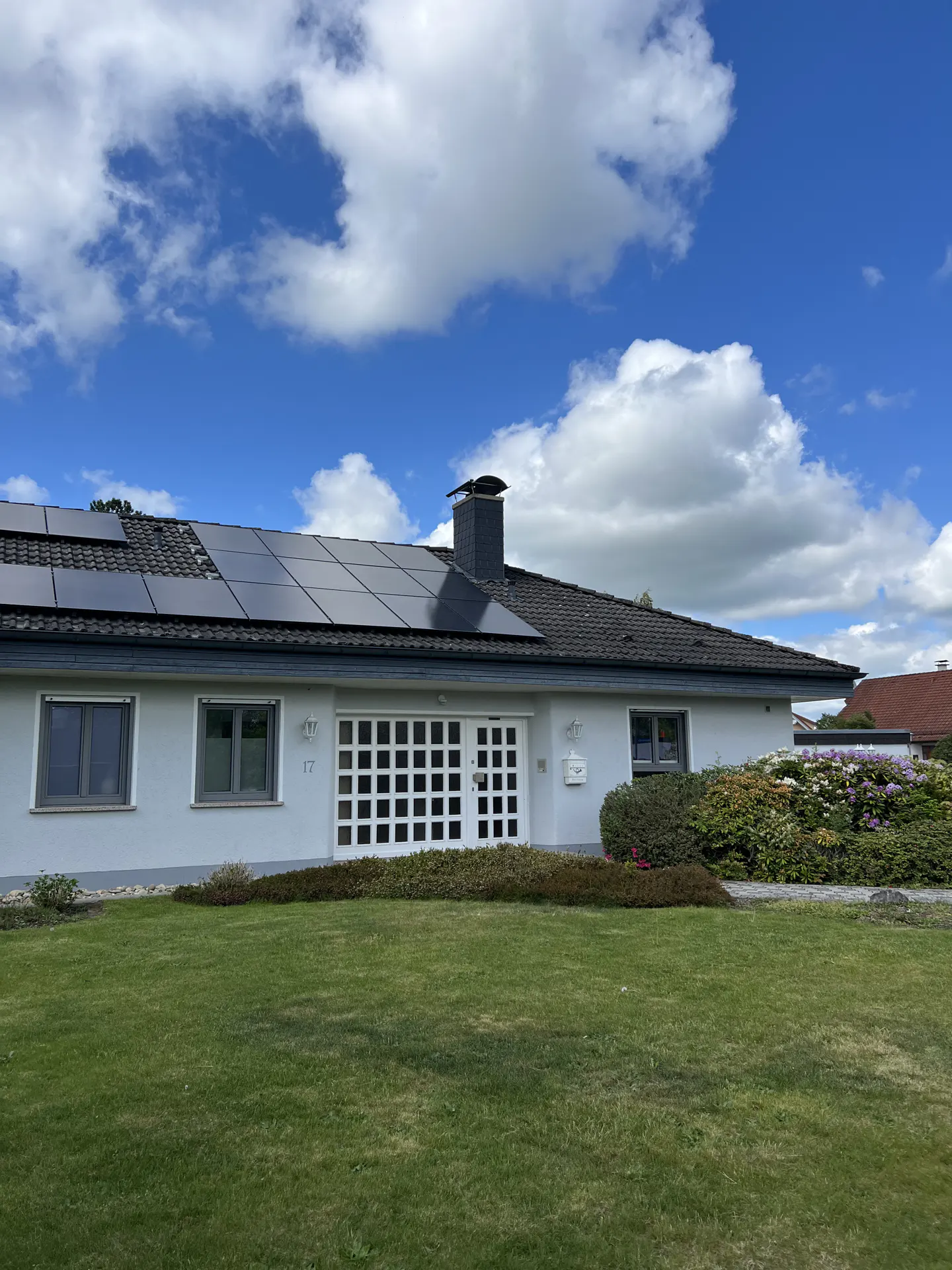 Single-story light gray house with solar panels on the roof, a grid-patterned front door, and a green lawn under blue sky.