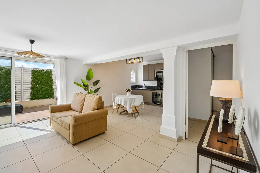 Bright, open-concept living space with a tan sofa, dining table, and kitchen. Sliding glass doors lead to a patio with green foliage.