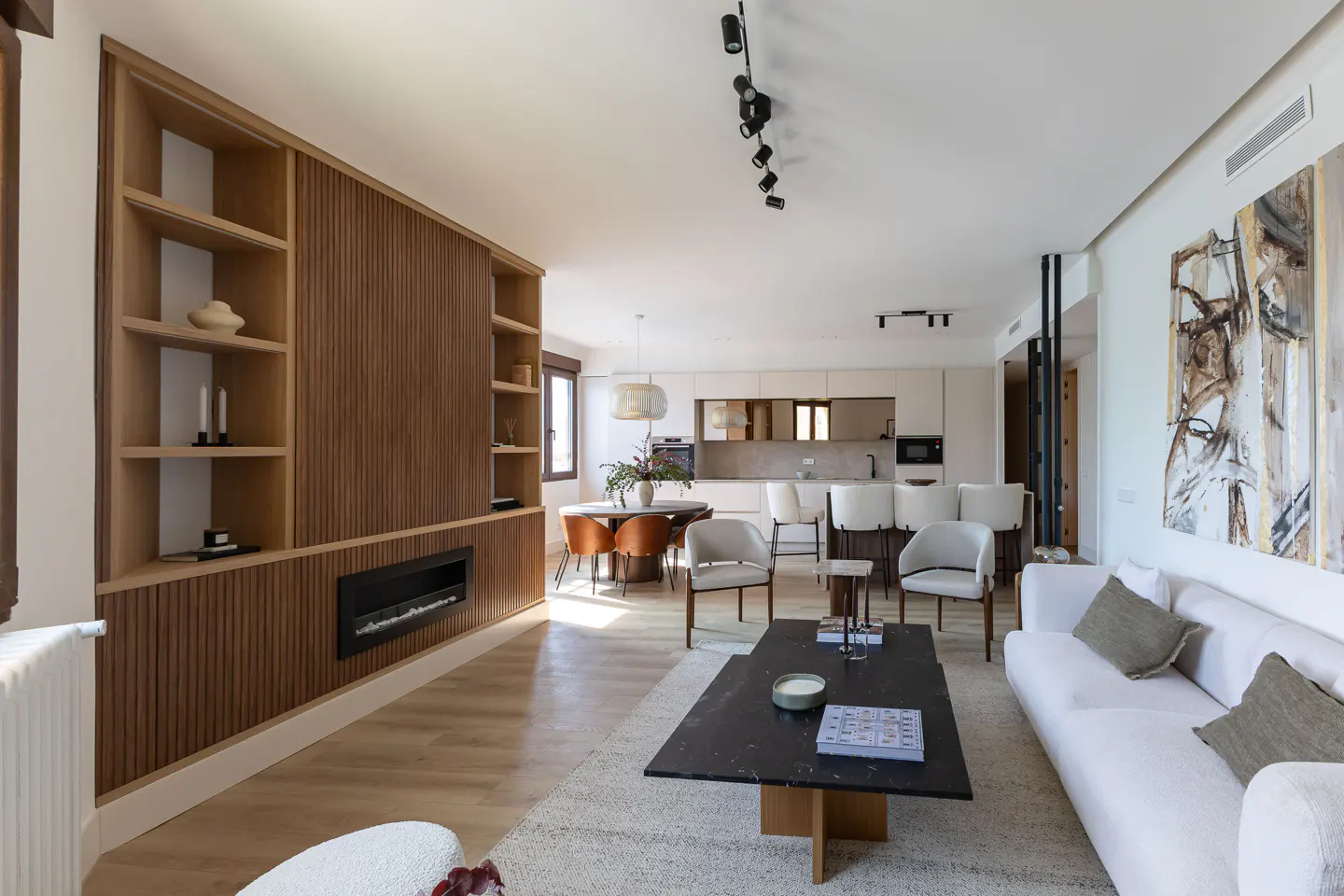 Bright, open-concept living space with a white sofa, black coffee table, and wood-paneled wall with shelves and a fireplace. A dining area and kitchen are visible in the background.