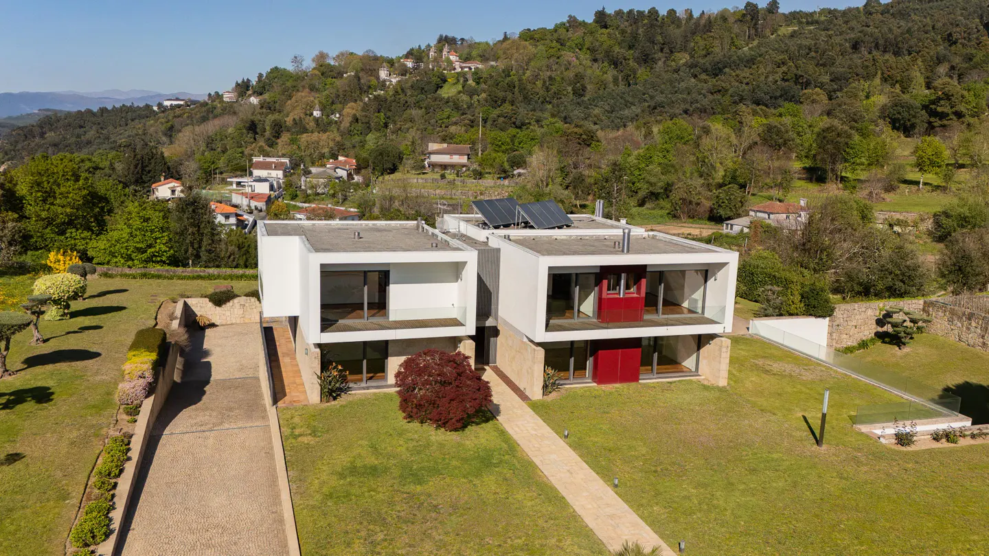 Aerial view of a modern, white, two-story house with red accents, solar panels, and a green lawn, set against a backdrop of lush, green hills.