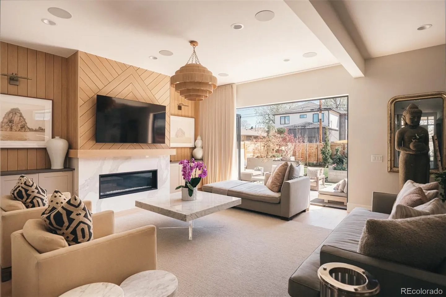 Living room with beige walls, wood paneling, and a marble fireplace. A large TV hangs above the fireplace. A tiered chandelier hangs from the ceiling.