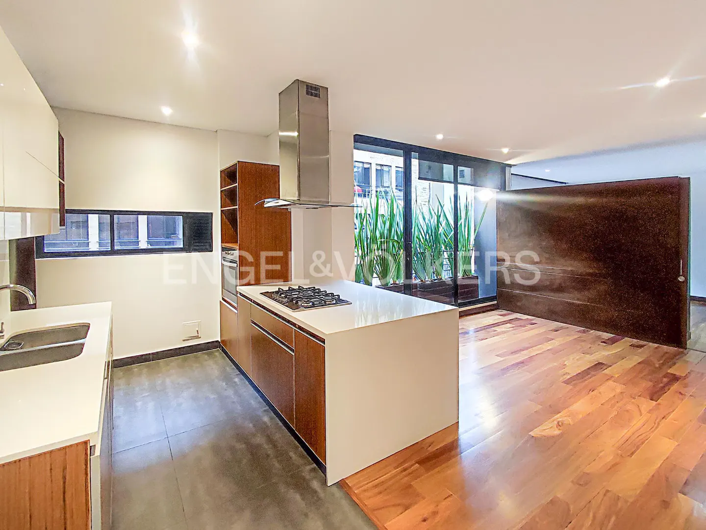 Open-concept kitchen with wood floors, white cabinets, and a large island with a cooktop. A window overlooks greenery.