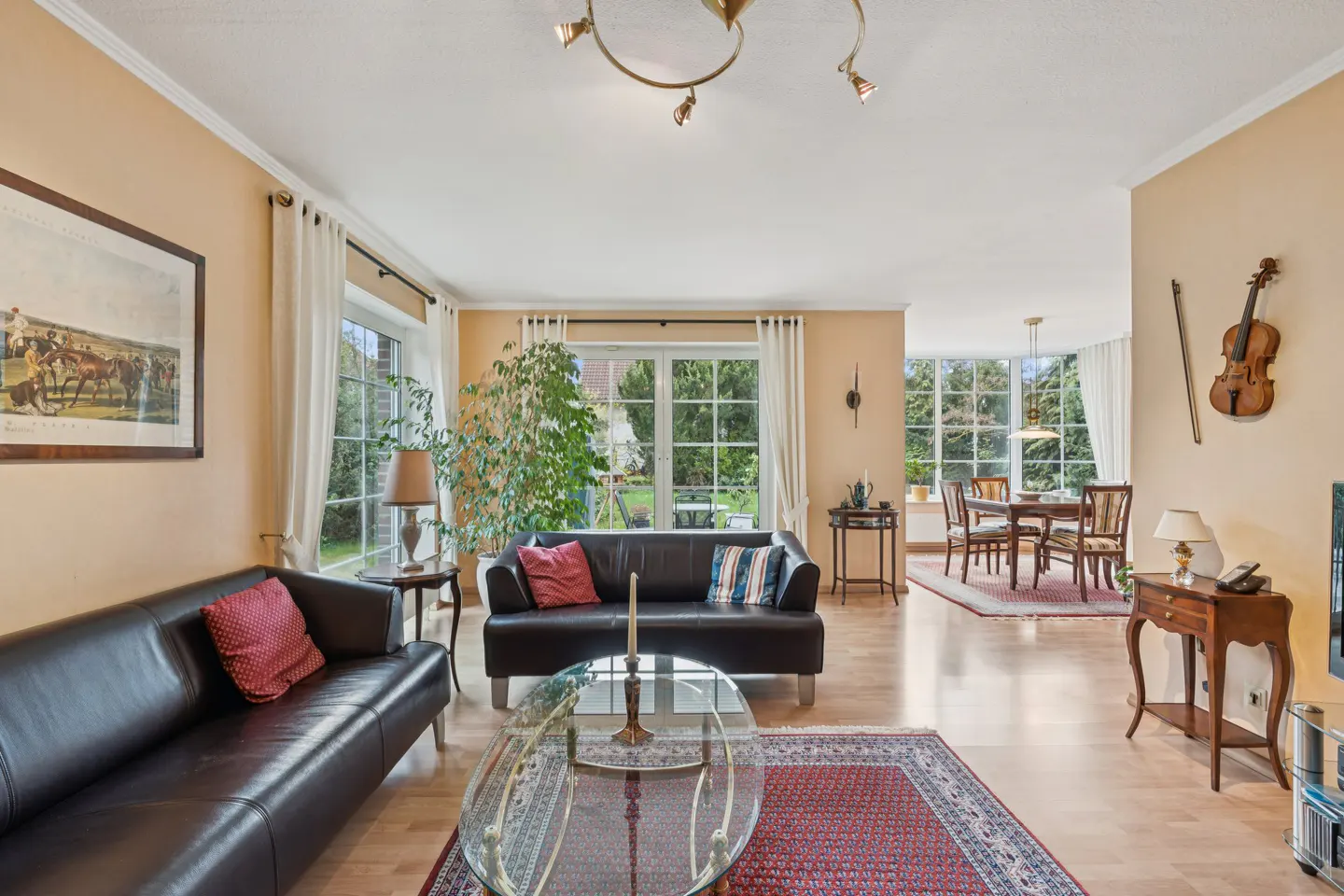 Living room with black leather sofas, glass table, and red rug. Large windows overlook a green yard. A violin hangs on the wall.