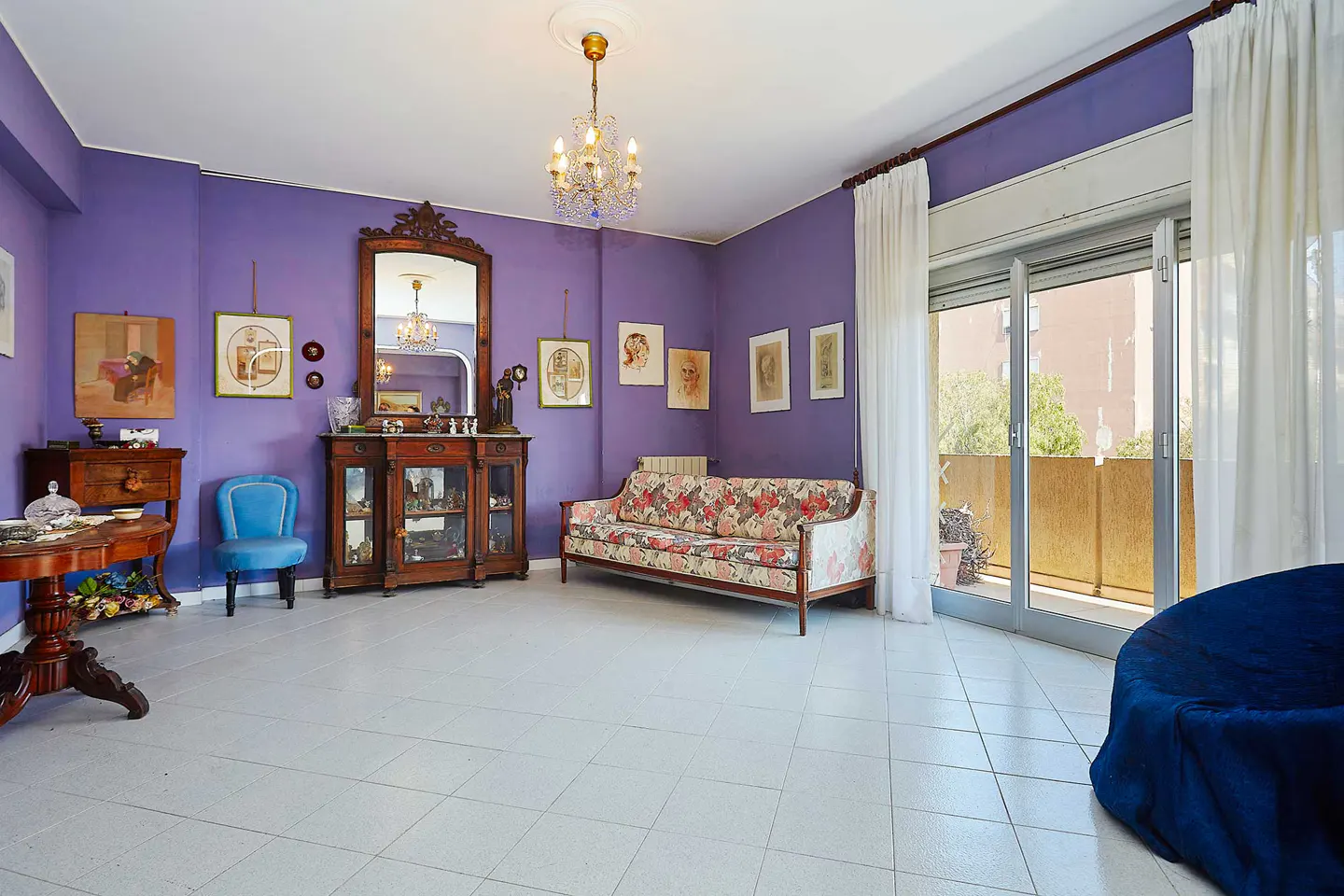 A living room with purple walls, white tile floors, and a floral sofa. A chandelier hangs from the ceiling. A balcony is visible through glass doors.