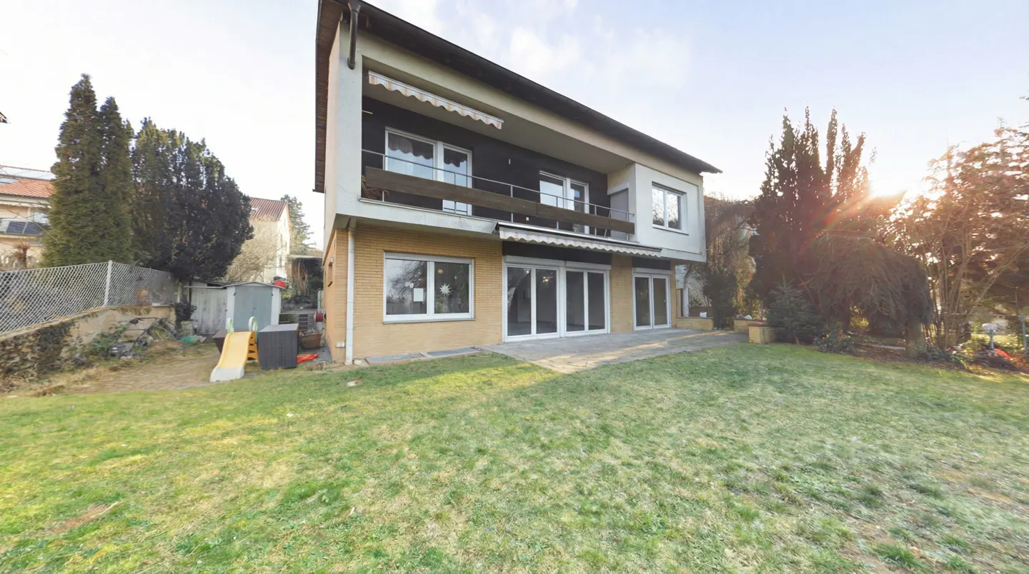Two-story house with a balcony and a large green lawn in the backyard. Trees and a blue sky are in the background.