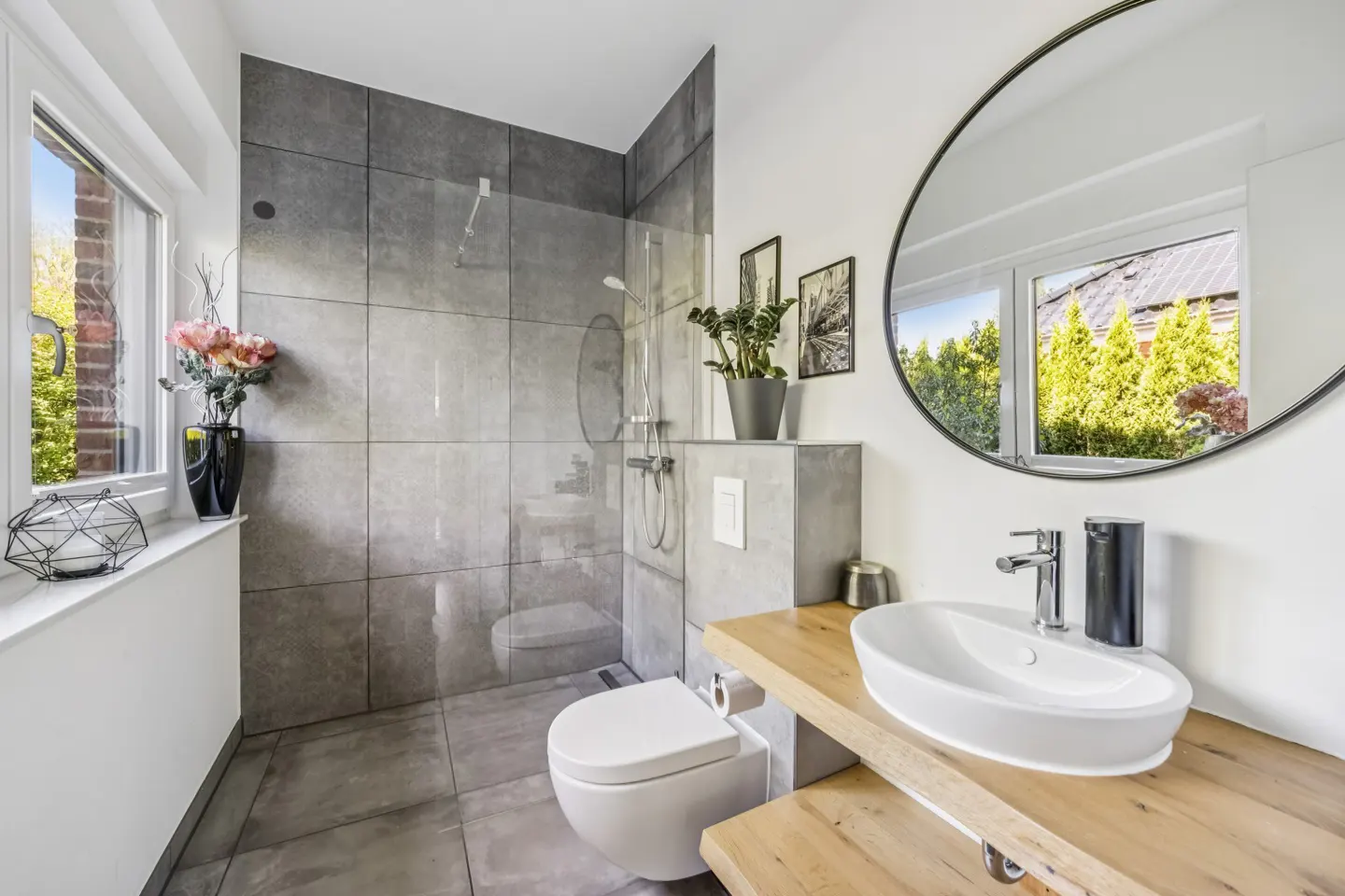 Modern bathroom with gray tile shower, white toilet, and wood countertop with oval sink. Round mirror reflects window view.