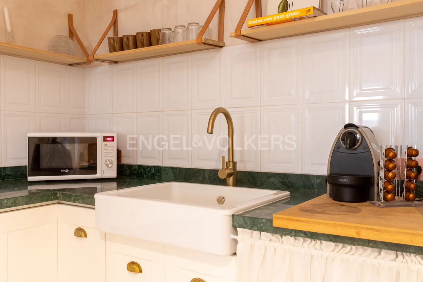 Modern kitchen with white farmhouse sink, brass faucet, green marble counters, microwave, and coffee machine on wood shelves.