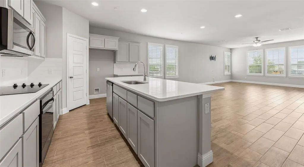 Open-concept kitchen and living room with gray cabinets, island, and tile flooring. Natural light streams through the windows.