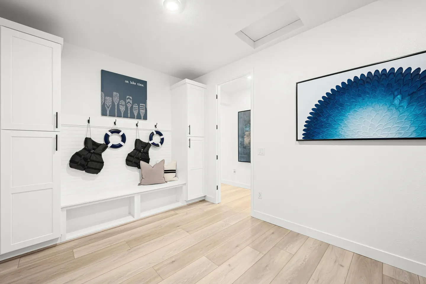 Bright mudroom with light wood floors, white walls, and white cabinets. Life vests and life preservers hang above a built-in bench. Blue artwork adorns the walls.