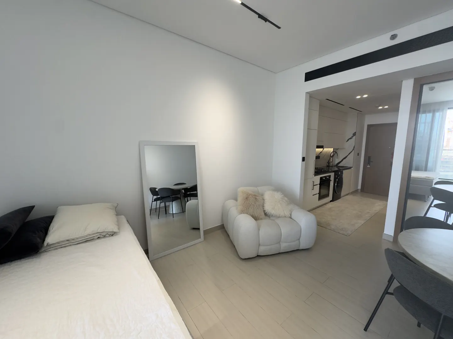 Bright studio apartment with a bed, white chair, and a mirror reflecting a dining table. A small kitchen is visible in the background.