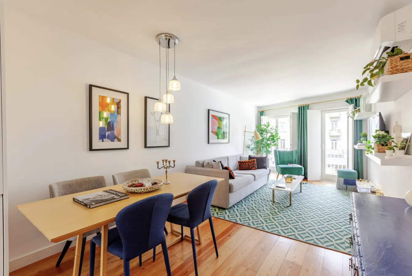 Bright living and dining area with wood floors, a light gray sofa, blue chairs, and green accents. Artwork adorns the white walls.