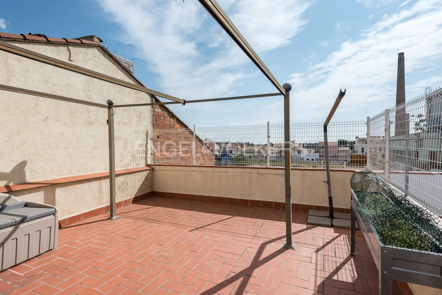 Rooftop terrace with red brick flooring, metal pergola frame, and white fence. A storage box and planter are visible. Cityscape in the background.