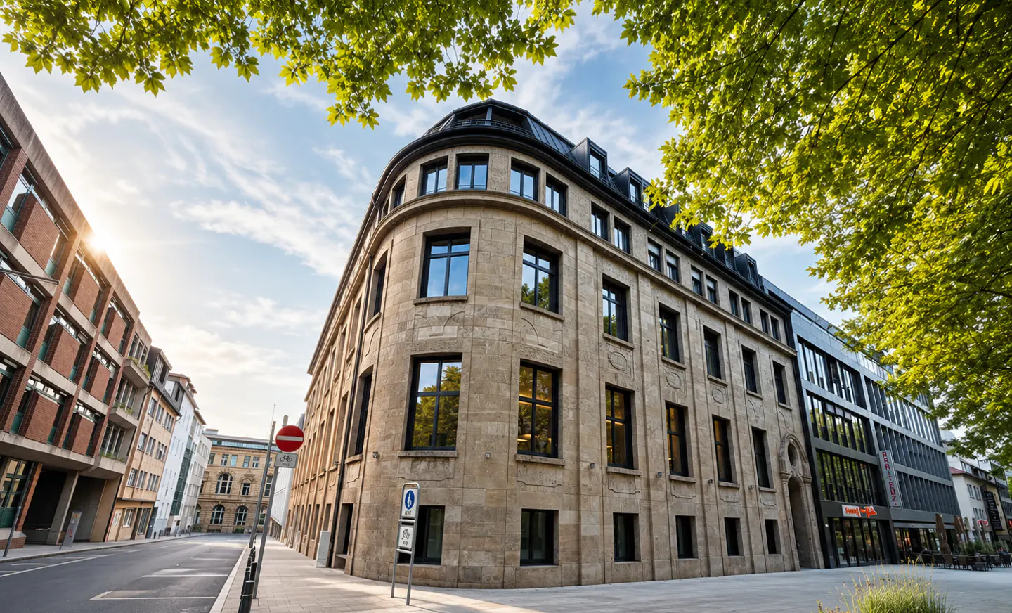 Street view of a tan stone building with black windows, framed by green trees and a blue sky.