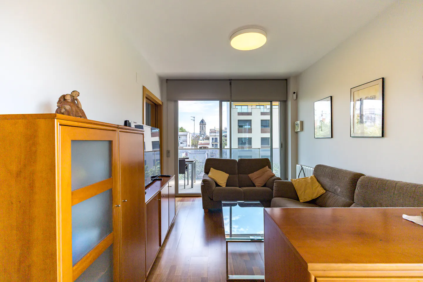 Bright living room with brown sofas, wood floors, and a balcony overlooking a city. A wooden cabinet stands against the wall.