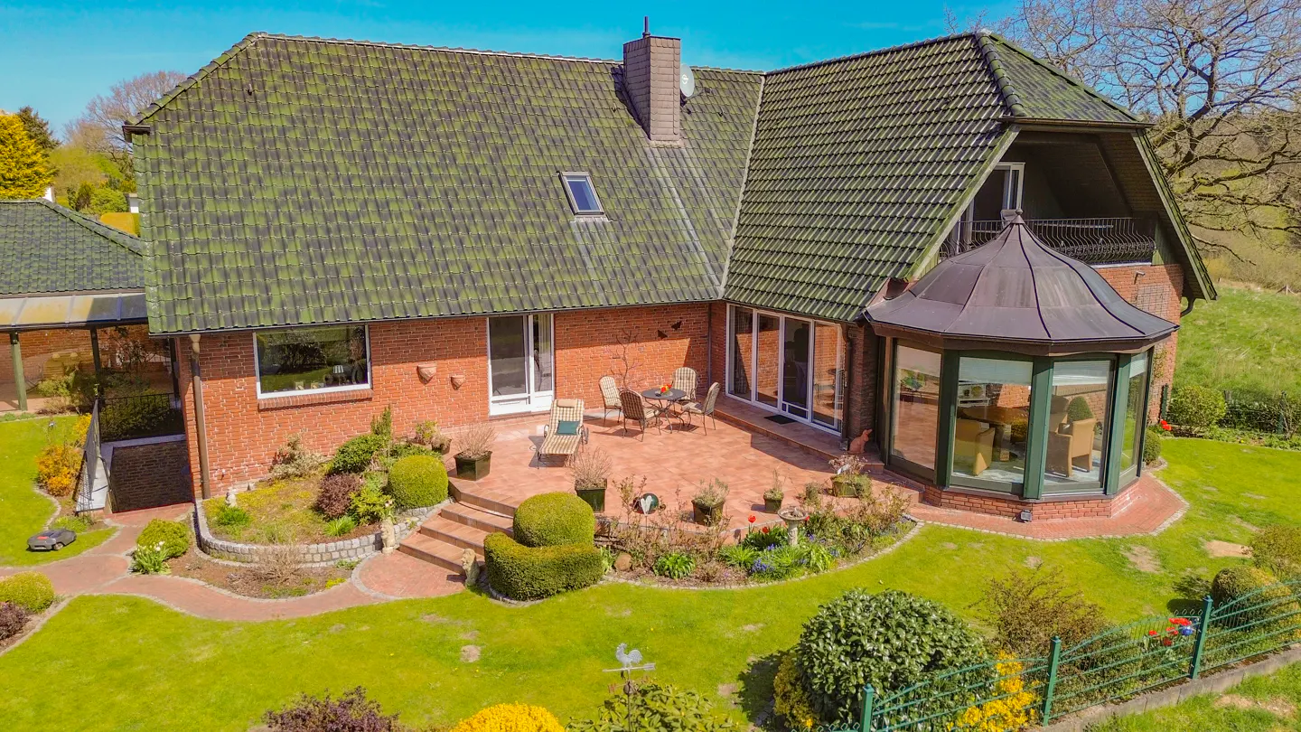 Aerial view of a red brick house with a green tile roof, a patio with furniture, and a well-manicured lawn.