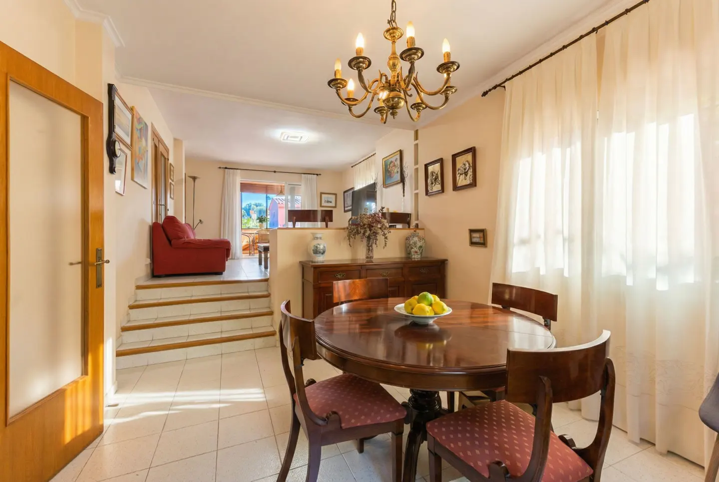 A bright dining room with a round wooden table, chairs, and a brass chandelier. A red sofa is visible in the background.