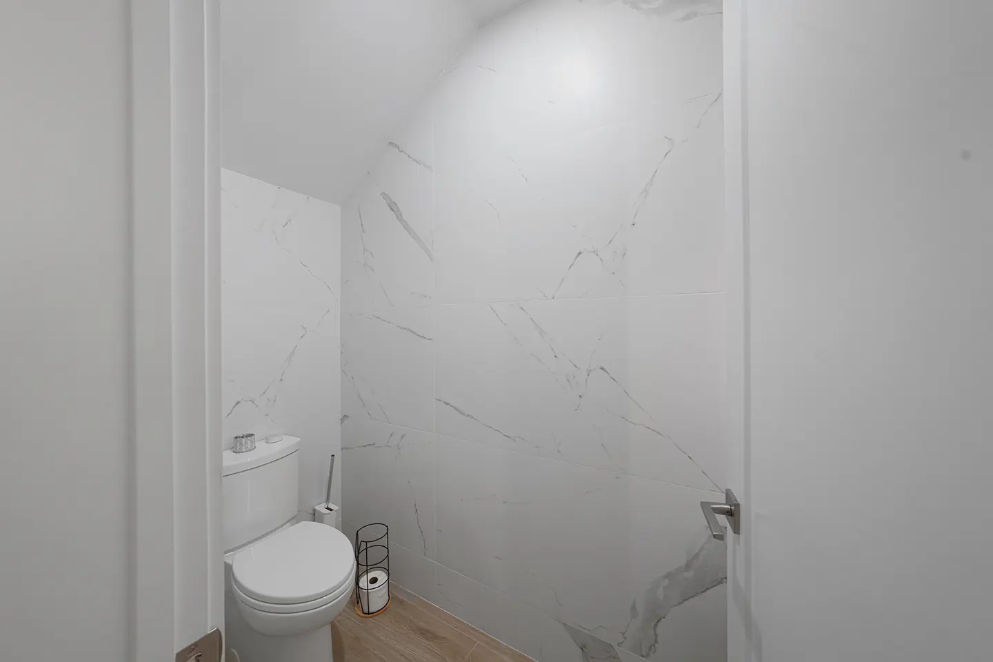 A small, modern bathroom with white marble walls, a toilet, and a wooden floor.