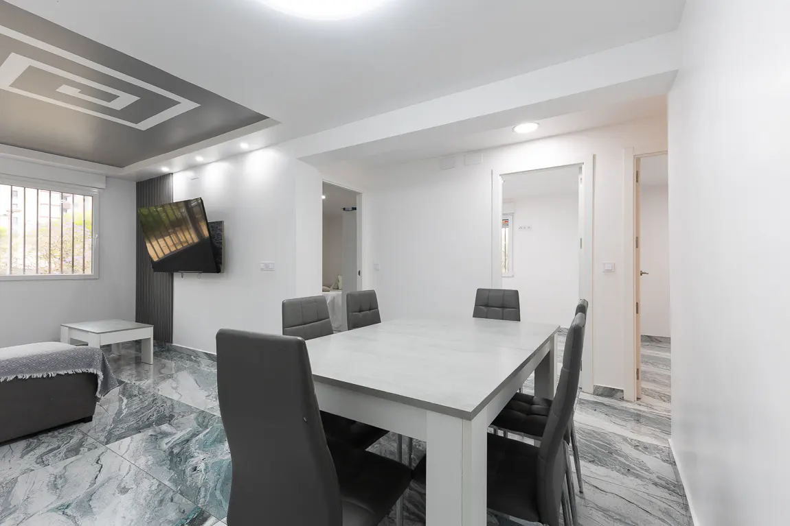 A modern dining room with a white table, gray chairs, and marble floors. A TV is mounted on the wall.
