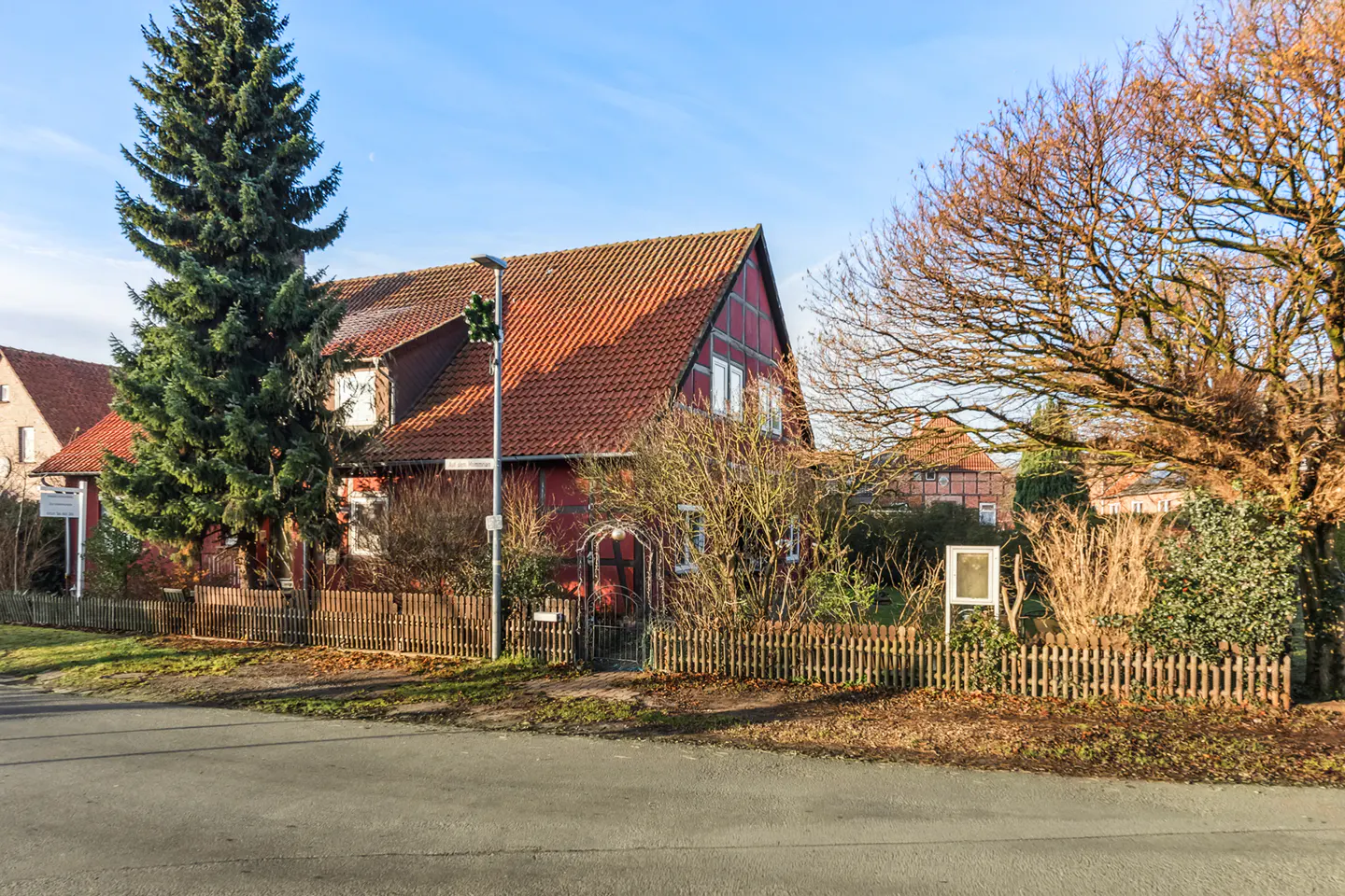 A red half-timbered house with a red tiled roof behind a wooden fence and a large pine tree.