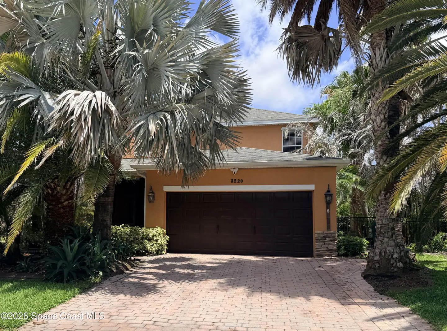 Two-story orange house with a brown garage door and brick driveway, framed by palm trees.