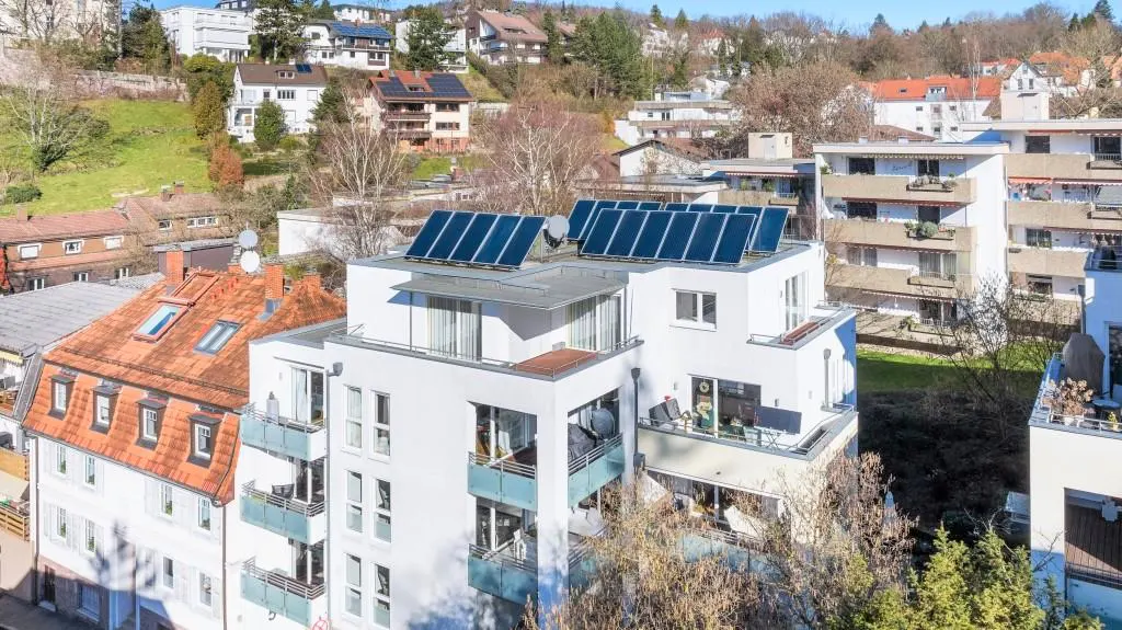 Aerial view of a modern white apartment building with solar panels on the roof, surrounded by other houses and trees.