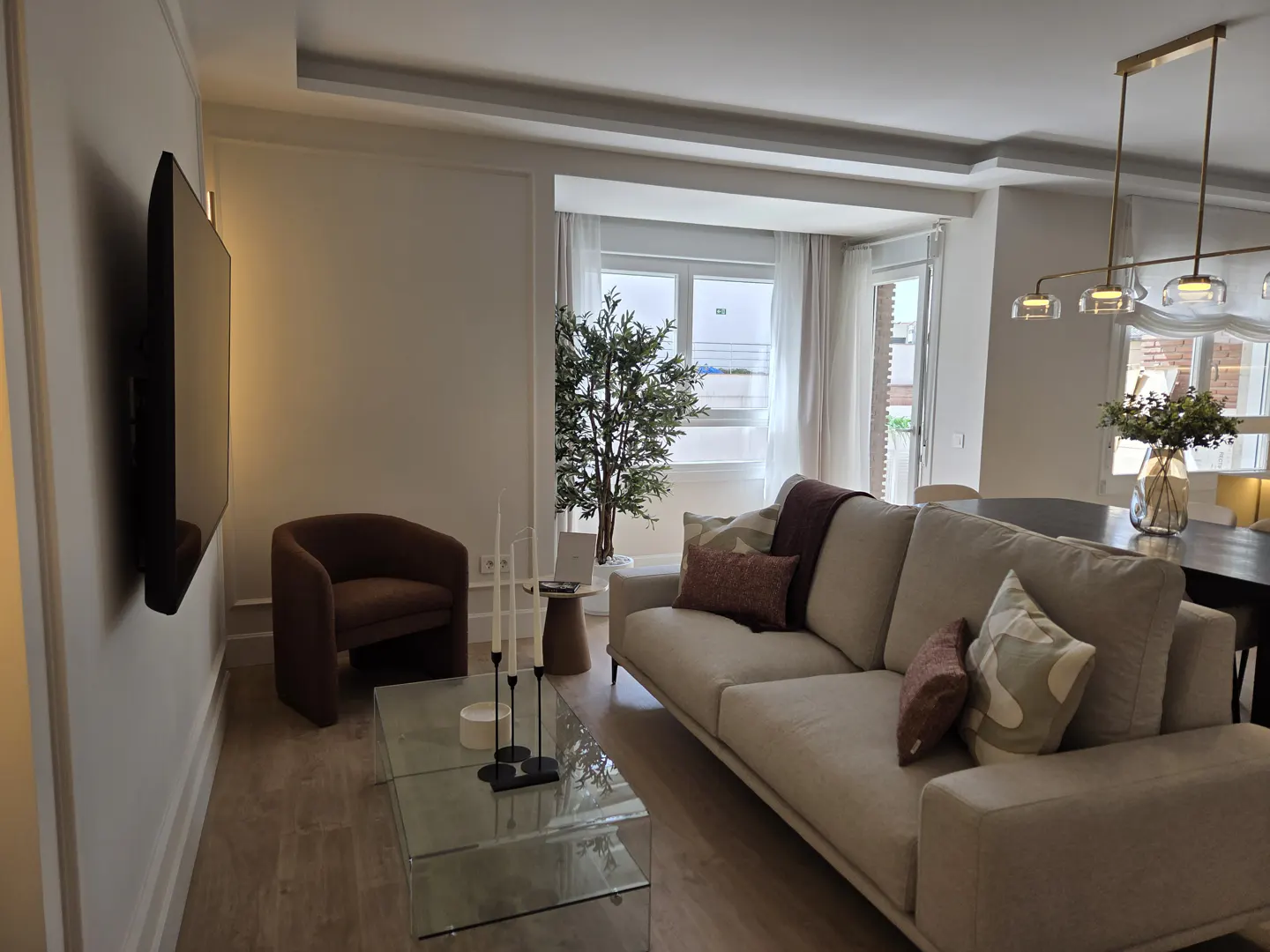 Modern living room with a beige sofa, brown armchair, glass coffee table, and a large window with a potted plant.