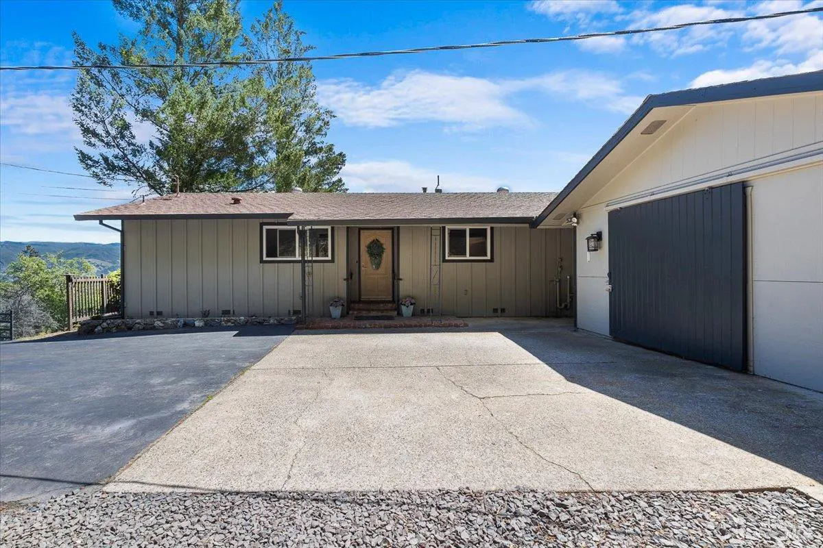 Exterior view of a one-story home with gray siding, a brown roof, and a large concrete driveway. A dark gray garage is attached to the right.