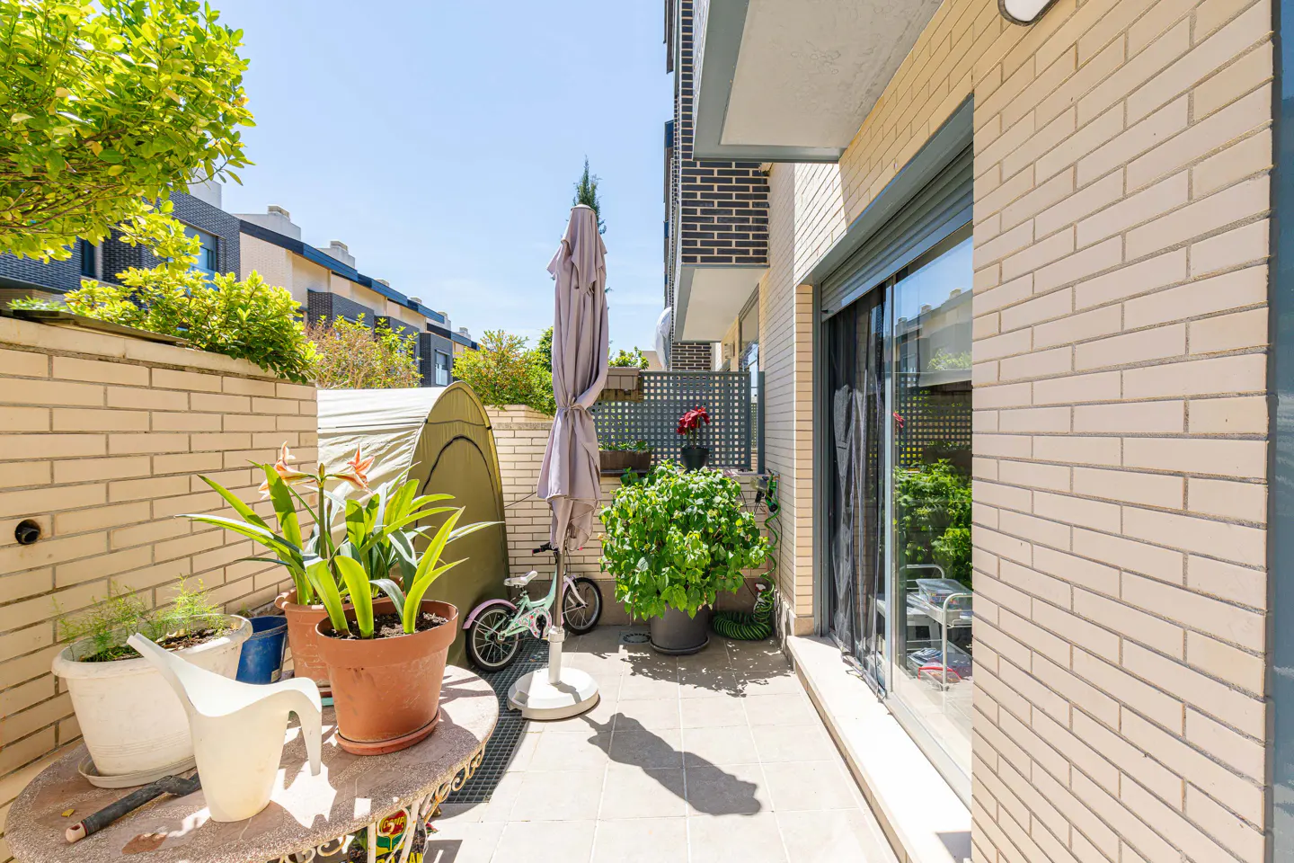 A sunny balcony with brick walls, potted plants, a small bike, and a closed gray umbrella. A tent is visible in the background.