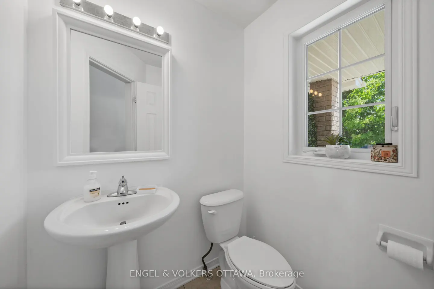 Bright white powder room featuring a pedestal sink, toilet, wall mirror, and a window with a view of green trees.