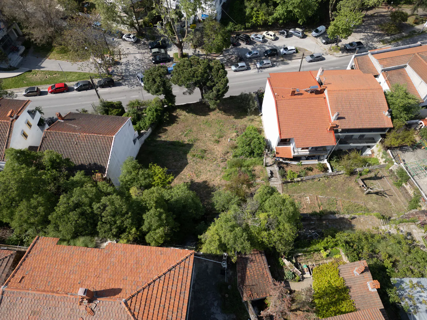 Aerial view of a vacant lot surrounded by houses with orange tile roofs and green trees. A street with parked cars is visible in the background.