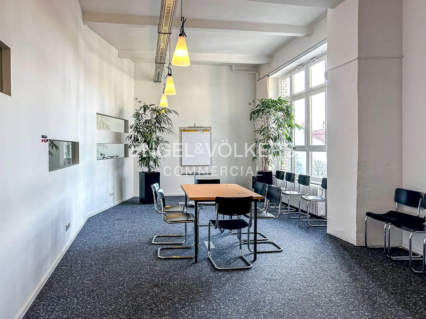 Bright office space with a table, chairs, whiteboard, and plants. The walls are white, and the carpet is gray.