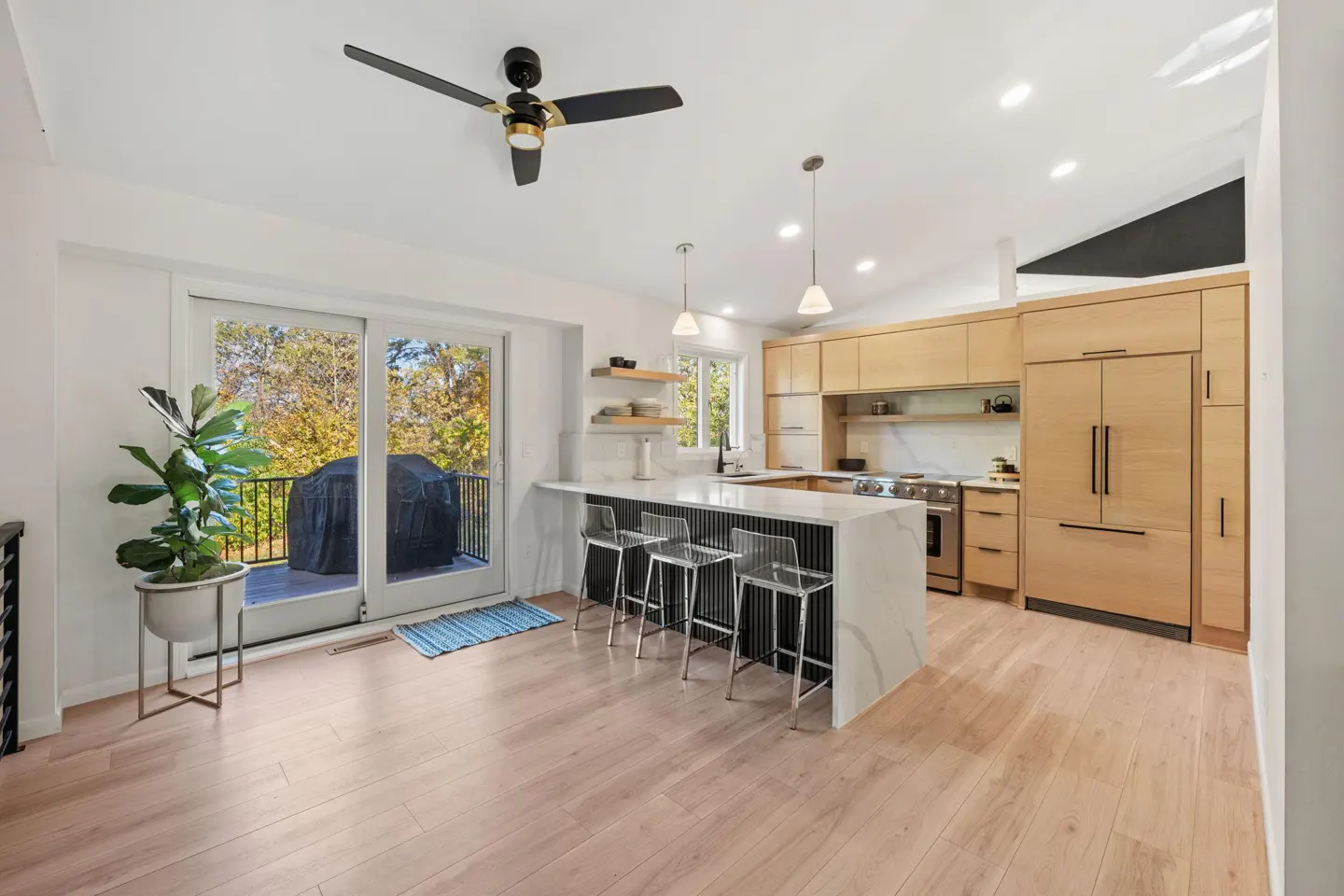 Bright, modern kitchen with light wood cabinets, white marble island with seating, and sliding glass doors to a deck. A black ceiling fan is visible.