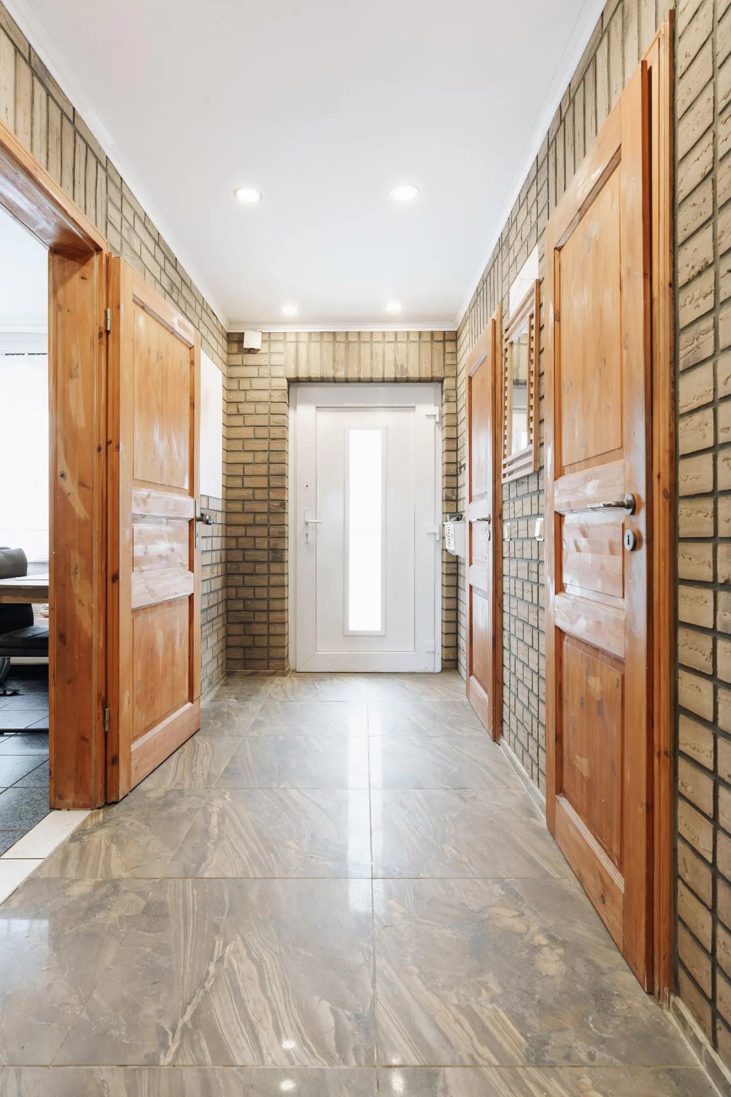 Hallway with brick walls, marble floors, and wooden doors. A white door is at the end of the hall.