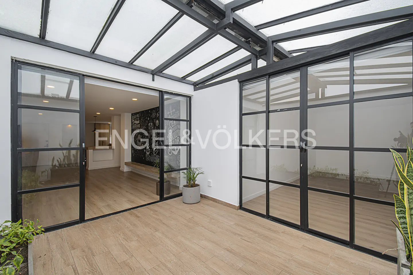 Sunroom with wood-look tile floor, black-framed glass doors, and a glass roof. The Engel & Völkers logo is visible.